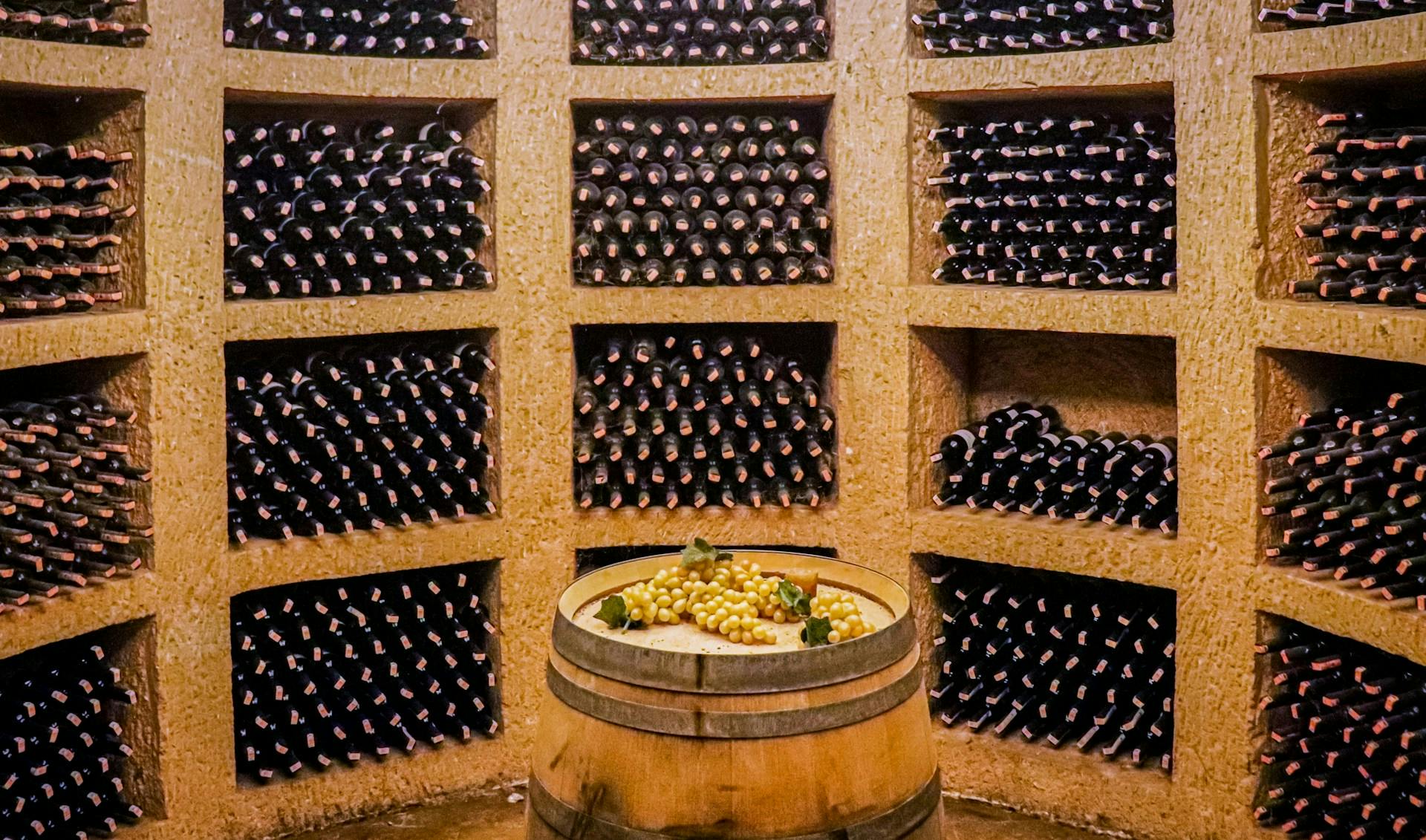 Weinregale voller Weinflaschen und ein Fass mit Weißtrauben in der Mitte