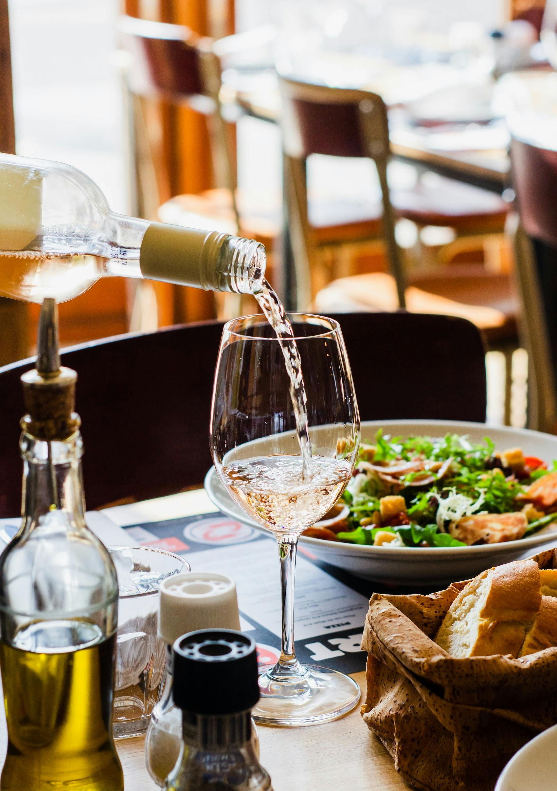 Klare Rose Flüssigkeit aus Weinflasche in Weinglas gekippt mit Essen im Hintergrund auf Holztisch