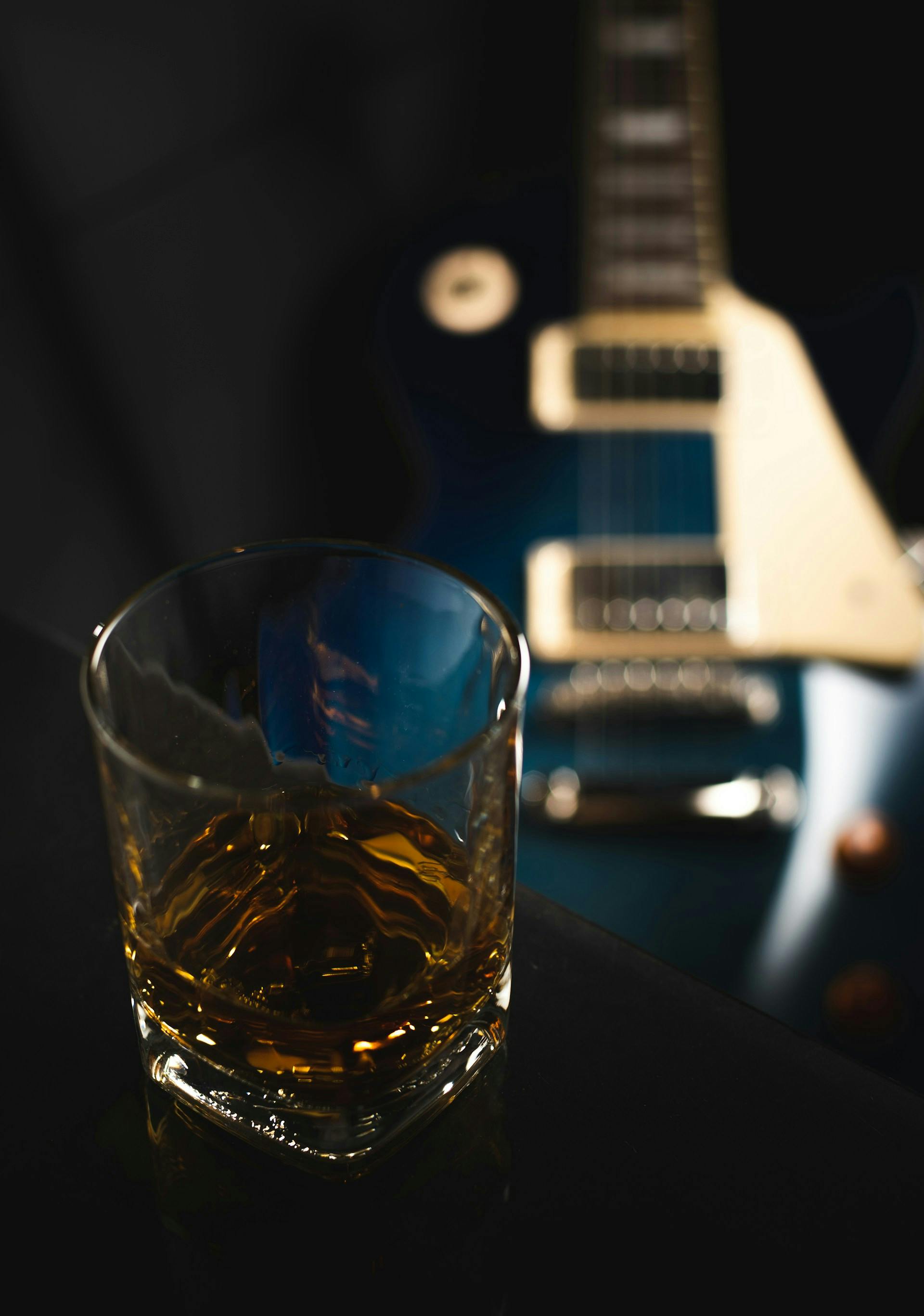 Glas mit brauner Flüssigkeit und E-Gitarre im Hintergrund