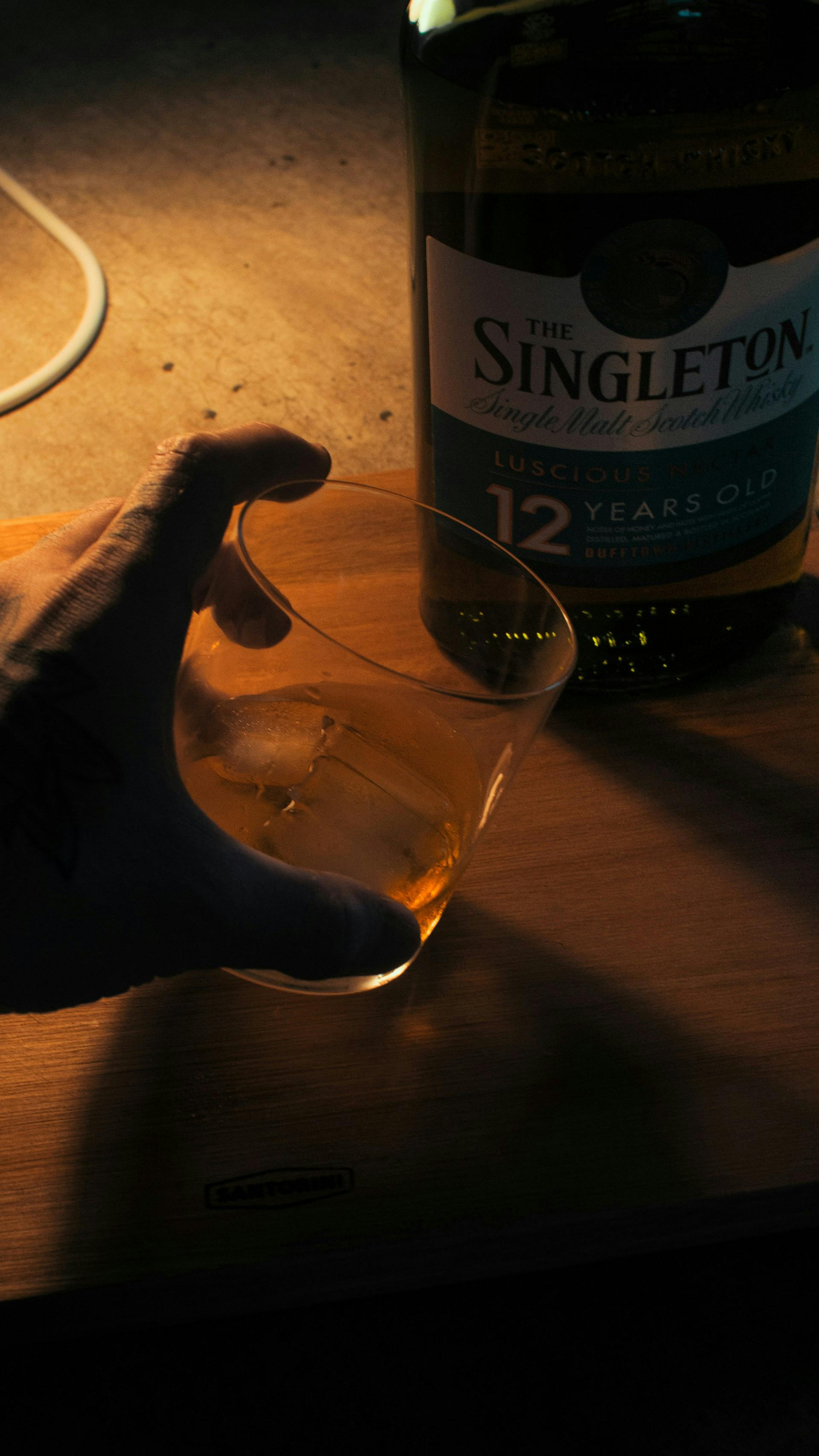 Flasche auf Holztisch mit Glas in einer Hand wo braune Flüssigkeit drinnen ist